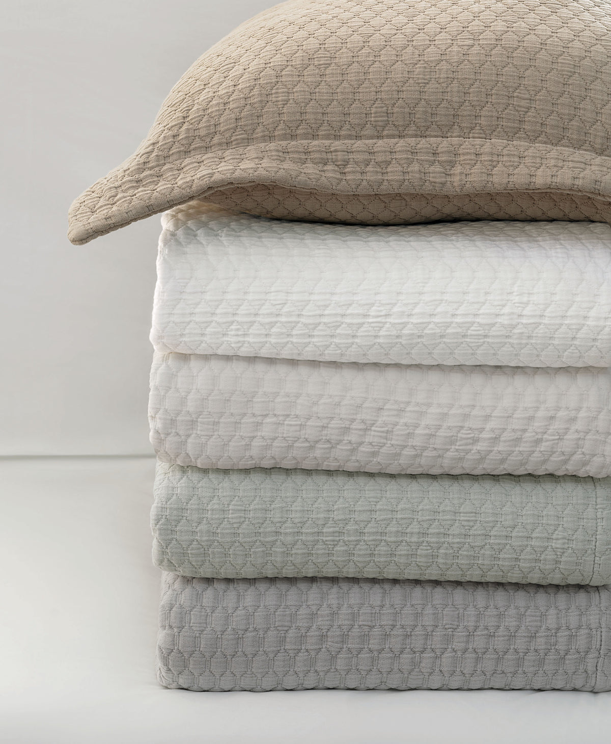 A stack of textured waffle-weave bedding, including a beige pillowcase and folded sheets in white, pale green, and light gray, set against a clean white background.