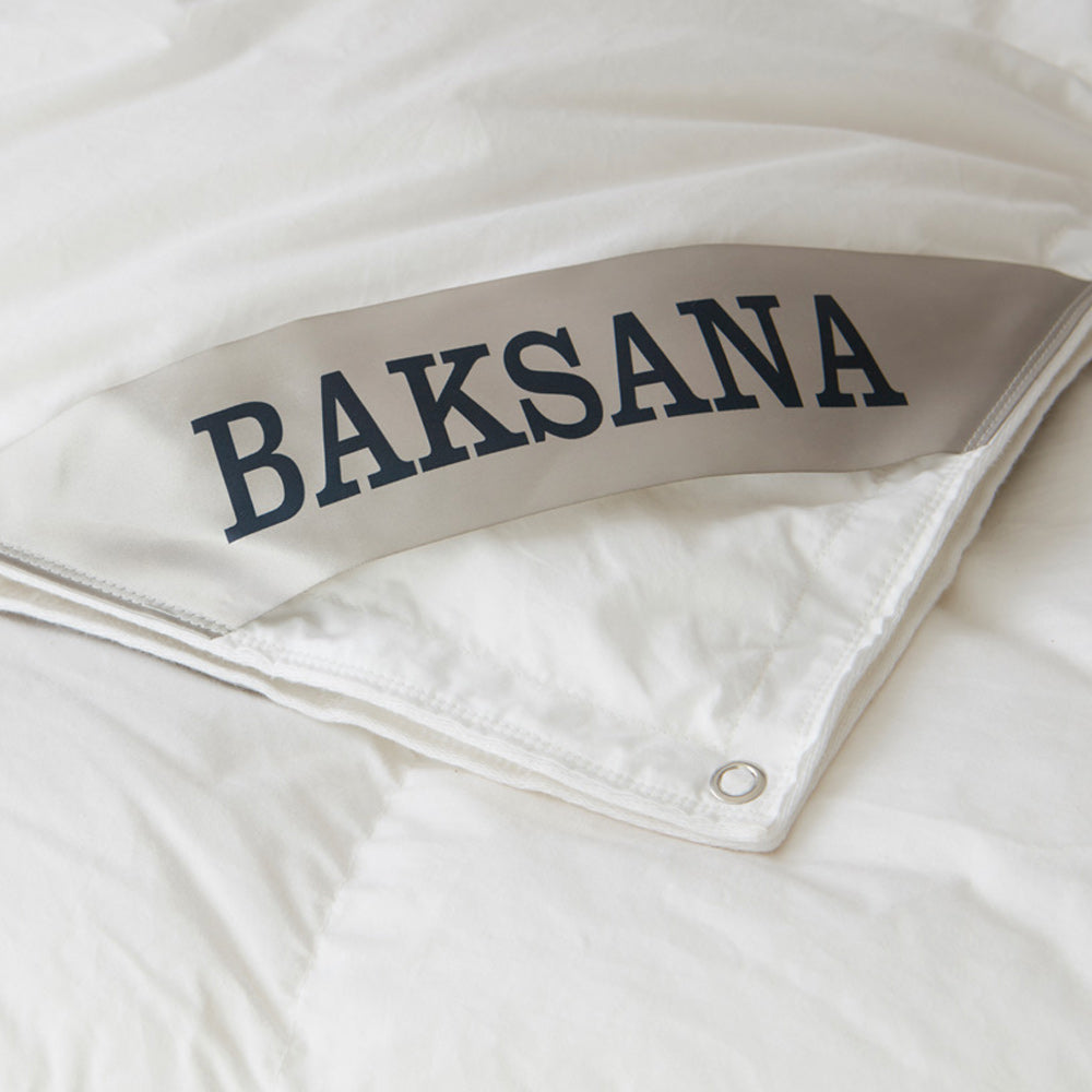 A close-up of a white quilted bedding item, likely a duvet cover or comforter, folded over to reveal a light gray band with the brand name 'BAKSANA' printed in dark navy lettering. A metal grommet is visible at the corner.