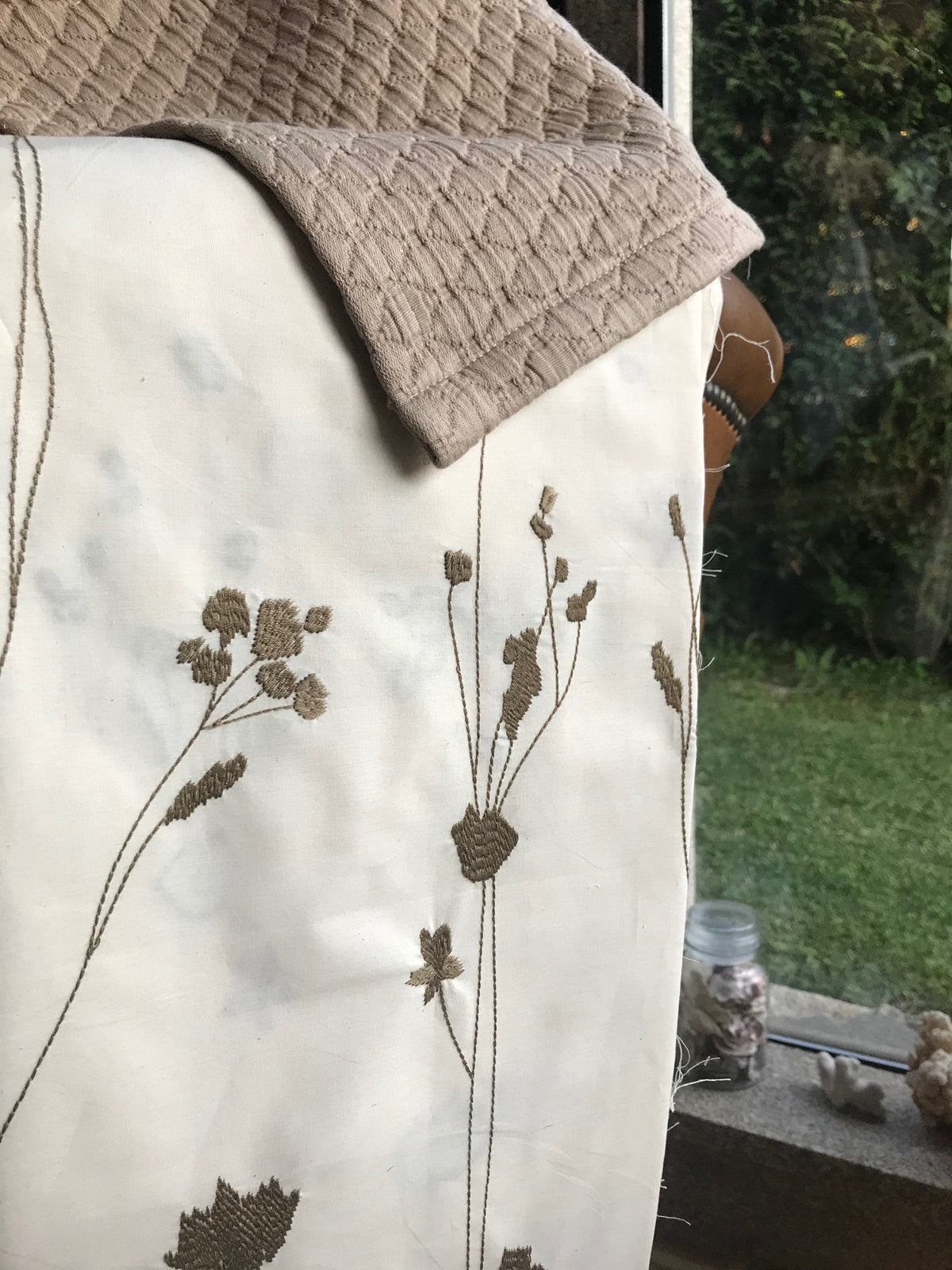 A close-up of a cream-colored fabric textile with olive embroidered wildflower motifs, draped over a surface next a window showing a green garden background.