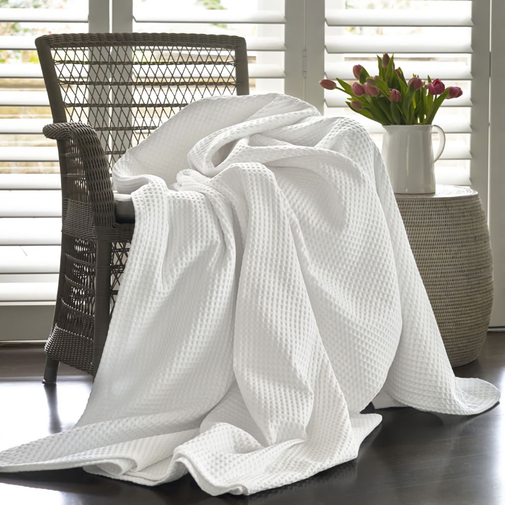 A white waffle-weave throw blanket is draped over a dark woven armchair in a bright room with white shutters, a side table, and a vase of pink tulips.