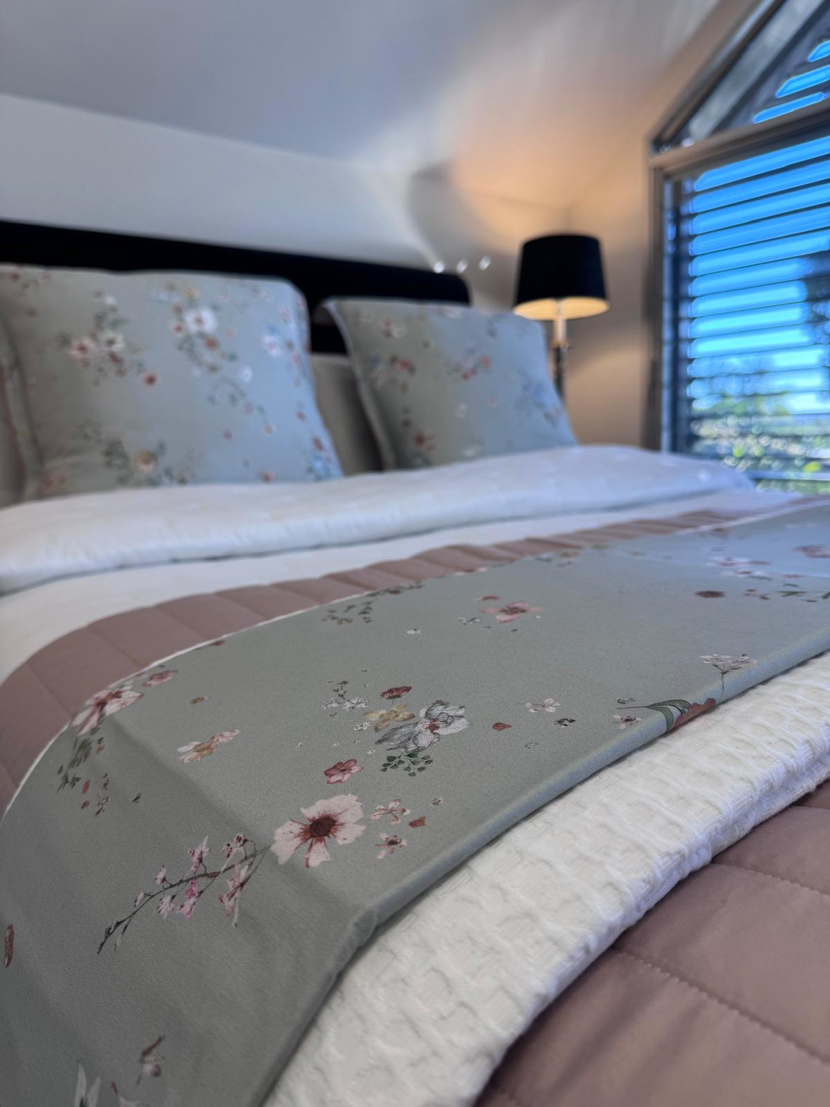 A close-up of a neatly made bed in a bedroom, featuring a sage green floral runner across the foot of the bed, white bedding, and matching floral pillows. A black lamp and window blinds are visible in the softly lit room.