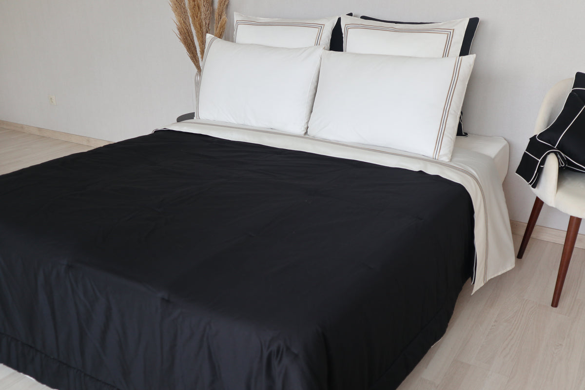 A neatly made bed dressed in a black bedspread called Glamorous with white pillows and white bedding called Golden visible at the top, photographed in a minimal bedroom setting with light wood flooring and a chair.