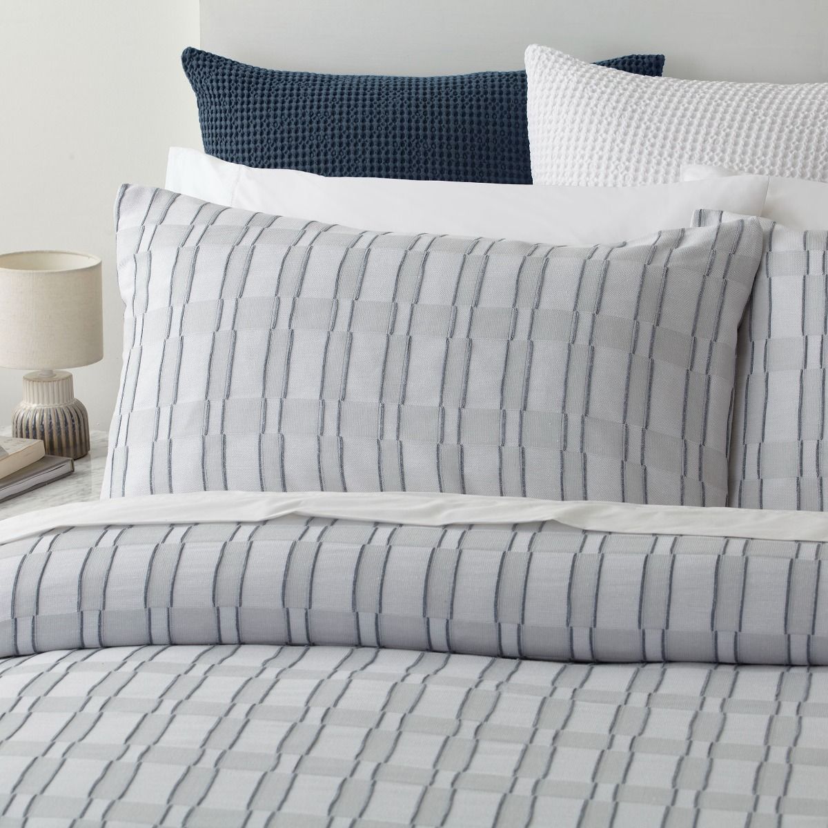 Denim Blue, Grey and white striped bedding set on a bed with pillows and a lamp in the background.