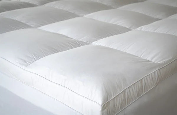 A close-up of a white quilted mattress topper or bed pad with padded square sections and stitched edges.