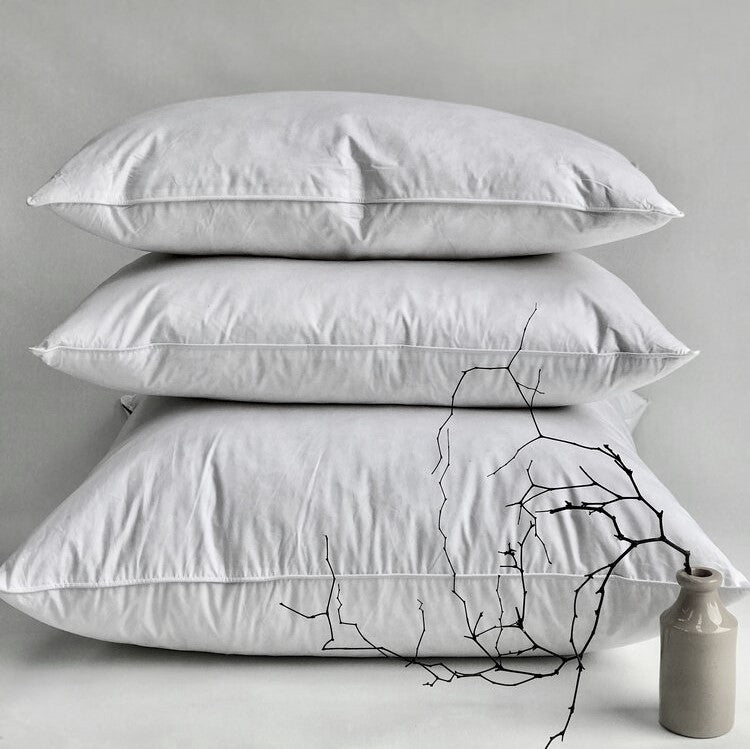 Three stacked white pillows on a light gray background with a small grey vase and black branch decoration in the foreground.