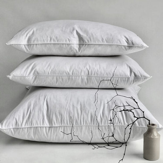 Three stacked white pillows on a light gray background with a small grey vase and black branch decoration in the foreground.