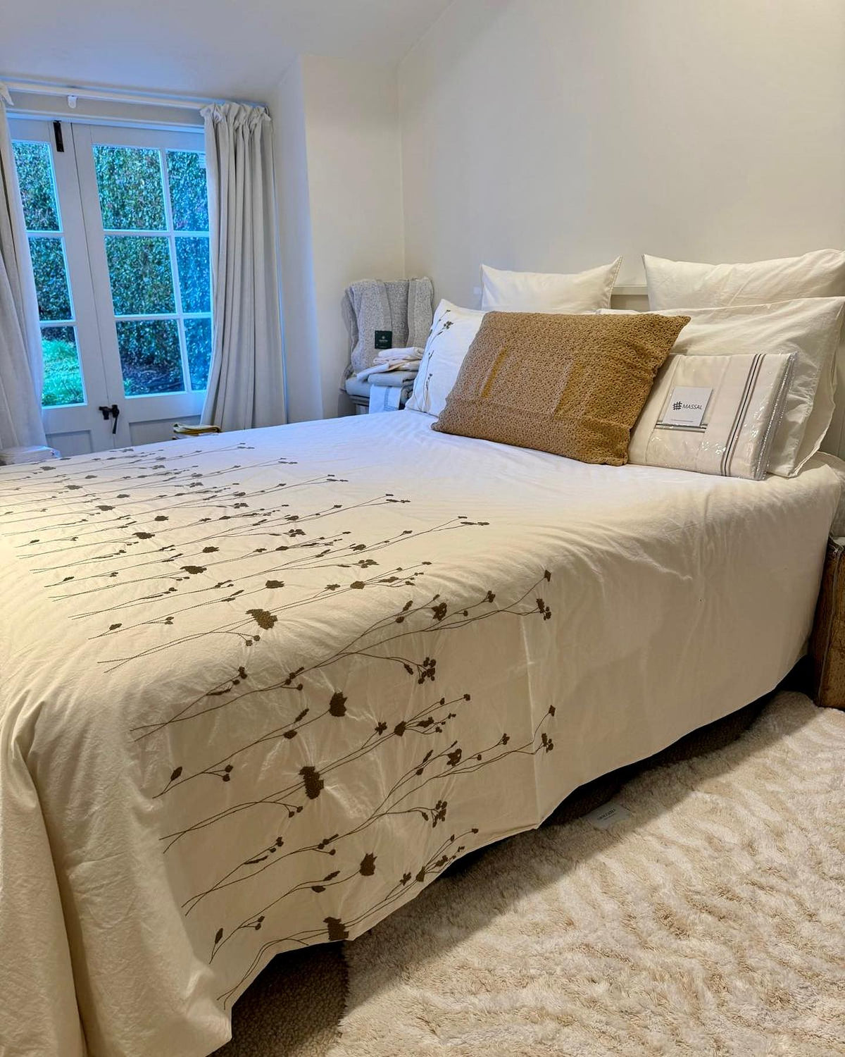A neatly made bed in a bright bedroom, dressed with an off-white duvet called Amalia by BOVI featuring a delicate olive floral branch pattern, white pillows, and a textured tan accent pillow.