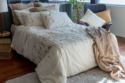 A styled bed in a bedroom setting featuring a cream-colored quilted duvet cover called the Amalia by BOVI with delicate olive floral embroidery, matching pillow shams, a textured beige throw blanket draped over the side, and a dark blue armchair near a window.