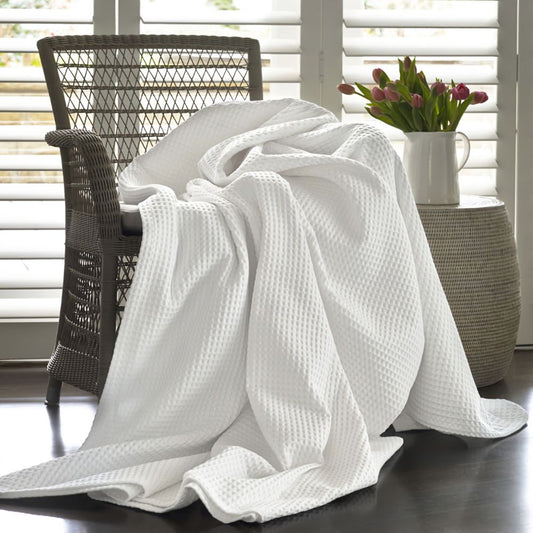 A white waffle-weave throw blanket is draped over a dark woven armchair in a bright room with white shutters, a side table, and a vase of pink tulips.