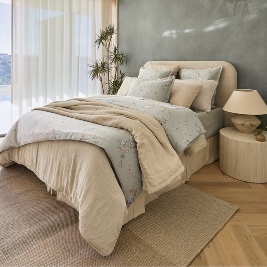 A styled bedroom scene showing a bed dressed in a light green floral bedding set layered with beige duvet and pillow shams, set against a textured gray wall and wood floor.