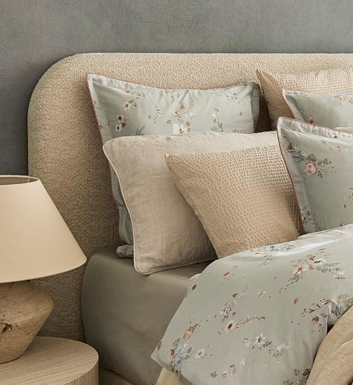 A styled bed with neutral-toned bedding and floral accent pillows, set against a textured beige upholstered headboard and a gray wall. A matching bedside table lamp is visible on the left.