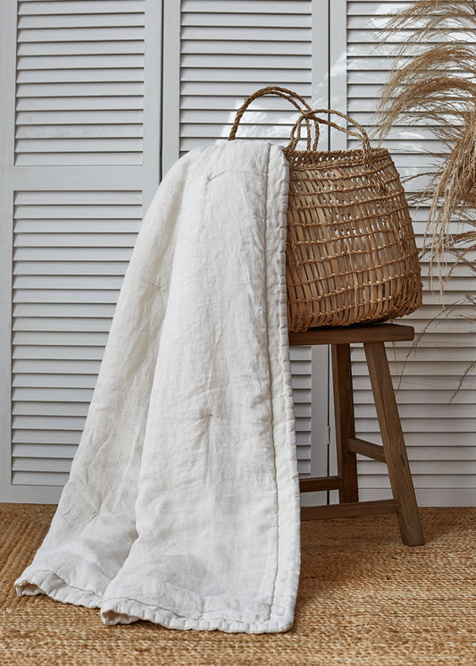 An off-white linen bedspread with a ruffled edge draped over a rustic wooden stool next to a woven straw tote bag, set against a white shutter backdrop.