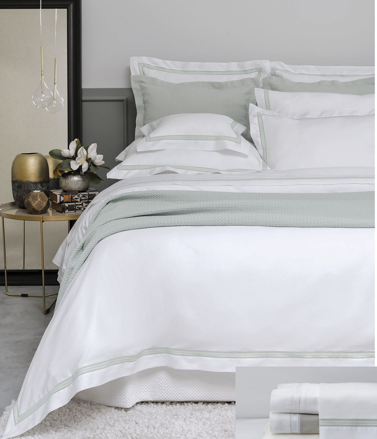 A neatly made bed dressed in white bedding with pale sage green striped accents and a matching sage green throw blanket, styled in a modern bedroom setting with a side table and mirror.