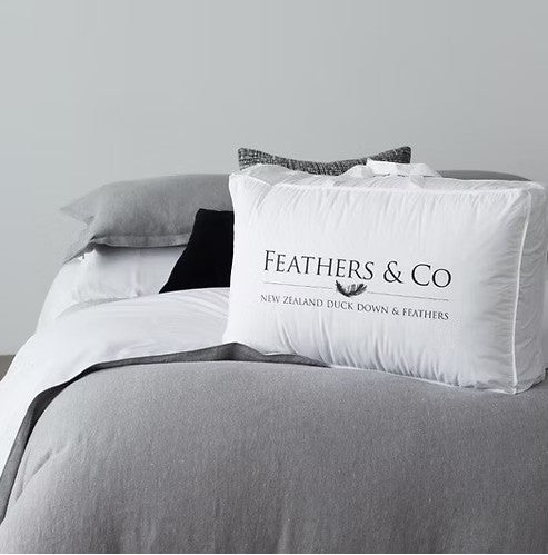 A white pillow from Feathers & Co, 30% Duck down & 70% feather which is  printed with 'NEW ZEALAND DUCK DOWN & FEATHERS' and a small bird silhouette, resting on a bed with geay bedding and white sheets.