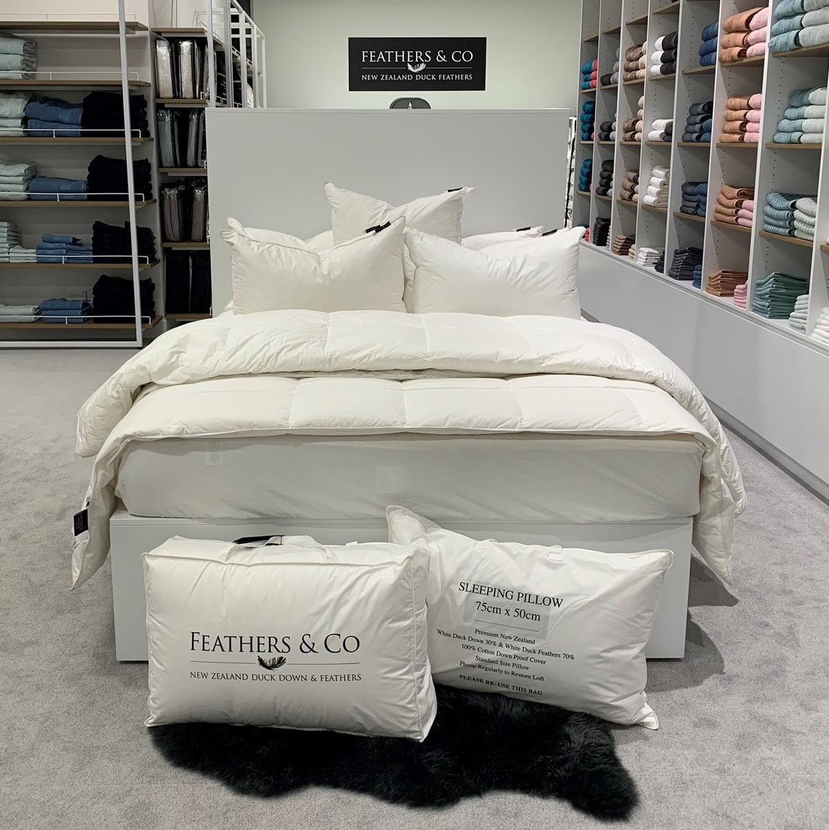 A bed dressed in a white quilted duvet and matching pillows, photographed in a bedding showroom. Two white pillowcases with printed text sit on the floor in front of the bed, and shelves of folded bedding are visible in the background.