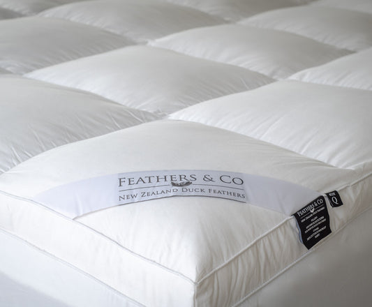 A close-up of a white quilted mattress topper with visible branding labels reading 'FEATHERS & CO' and 'NEW ZEALAND DUCK FEATHERS'.
