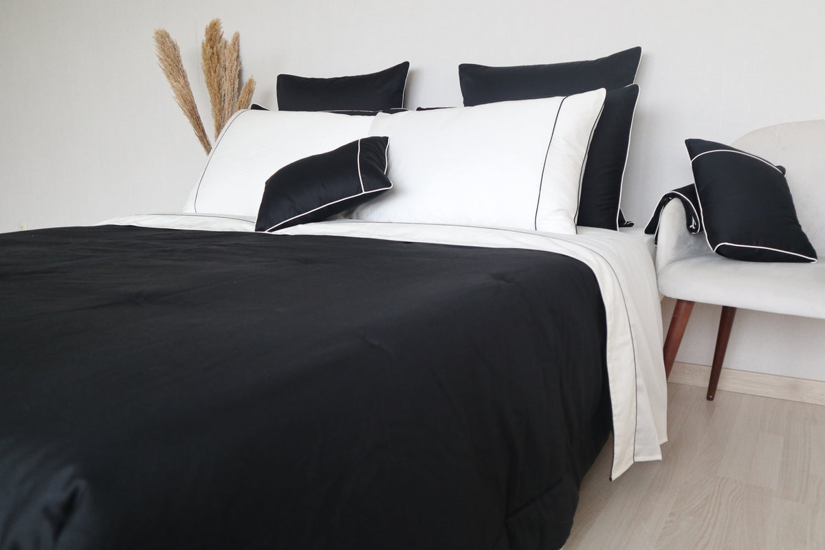 A bed dressed in a black and white bedding set called Glamorous by BOVI with multiple pillows, including two black pillows with white piping, set in a minimalist bedroom next to a white chair.