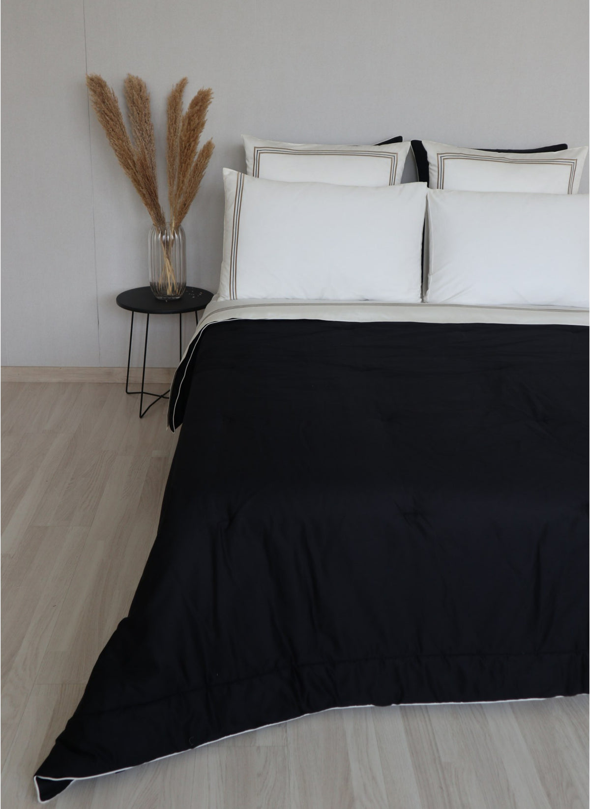 A bed dressed in a black duvet cover  called Glamorous with white piping trim, styled with white pillows in a minimalist bedroom setting with a small side table and dried pampas grass.