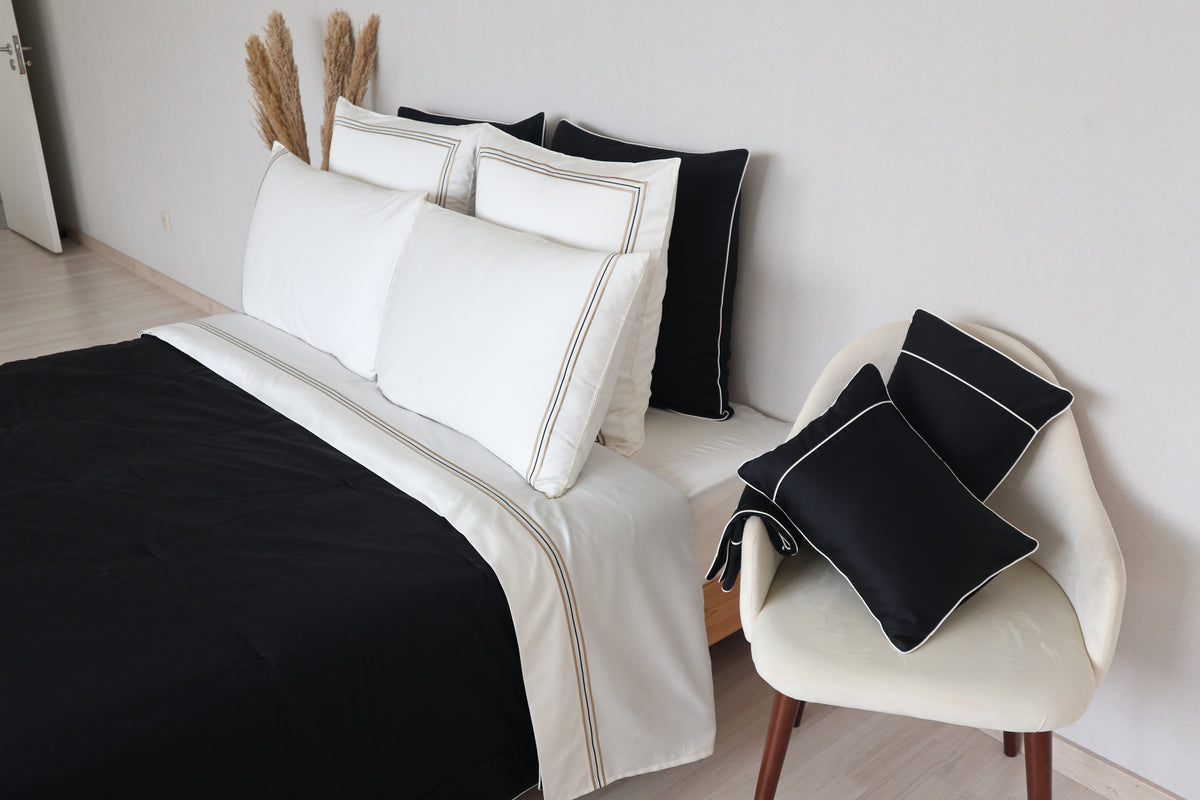 A styled bed with white bedding sheets called Golden and black pillows and a duvet cover called Glamorous, alongside a cream chair holding two black accent pillows called Glamorous with white piping, set against a light gray wall.