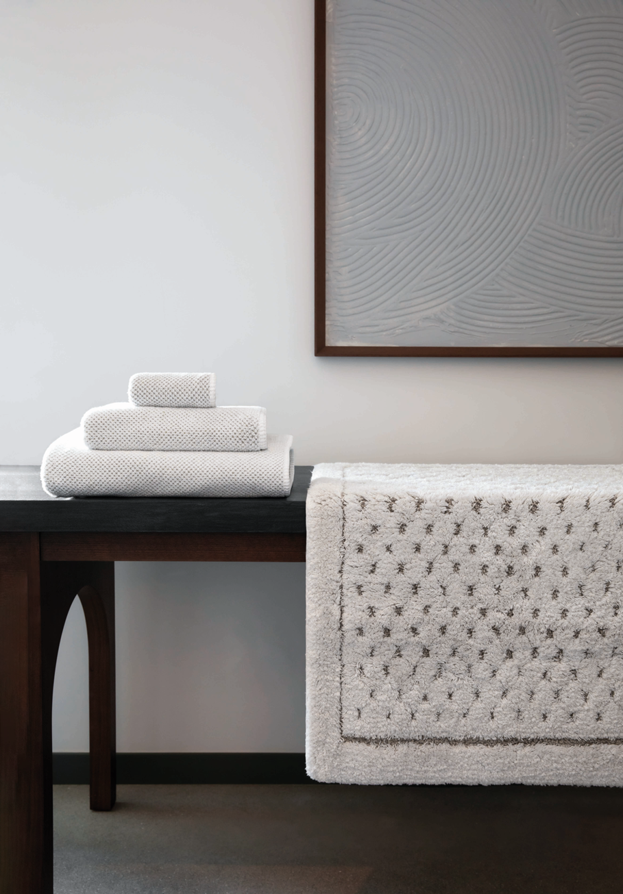 A stack of three folded white towels sits on a dark wooden table next to a textured off-white bath rug draped over the side of the table, with a minimalist framed wall art piece in the background.