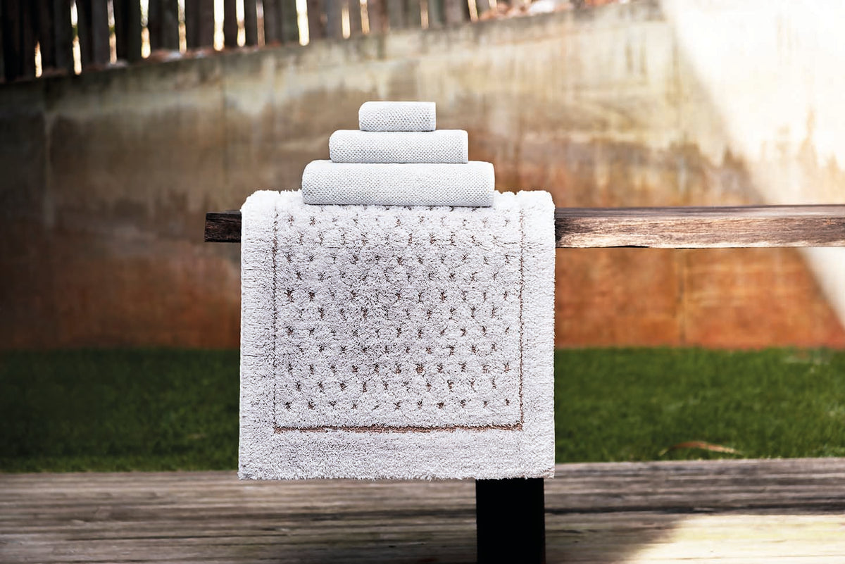 A set of four white textured bath towels, including a large bath rug draped over a wooden bench and three smaller towels stacked on top, set against an outdoor backyard background with grass and a wooden fence.