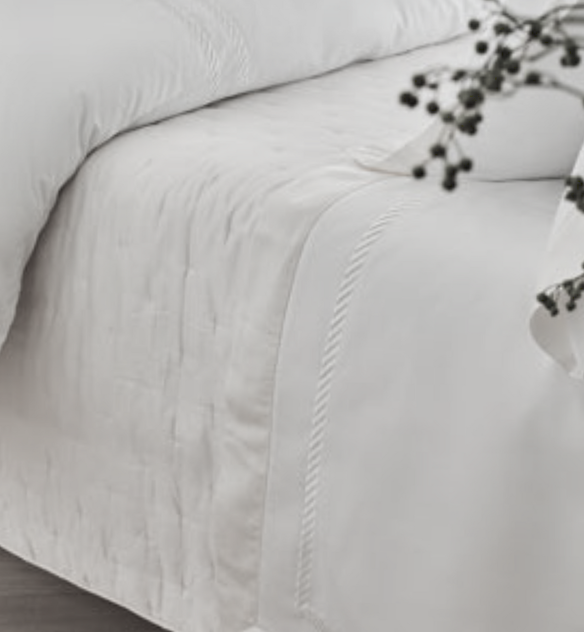 White quilted bedding set on a bed, featuring a textured quilted cover with a decorative braided edge and matching white pillows, styled with a delicate sprig of dark berries.