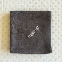 A dark gray textured makeup remover cloth with 'make-up' embroidered in white on the front, laid flat on a light beige textured surface.