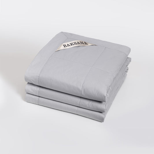 A folded light grey quilted coverlet called Luxury Espresso coverlet with a small cream fabric tag reading 'BAKSANA' on a clean white studio background.